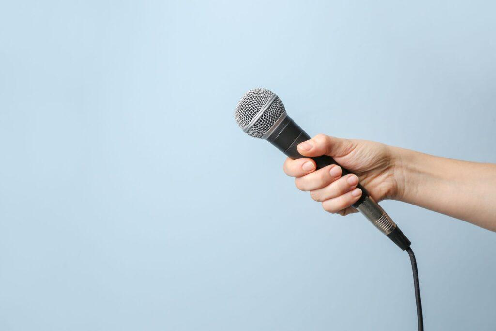 Nahaufnahme einer Hand, die ein Mikrofon vor einem hellblauen, neutralen Hintergrund hält, bereit für ein Interview oder eine Ansprache.
