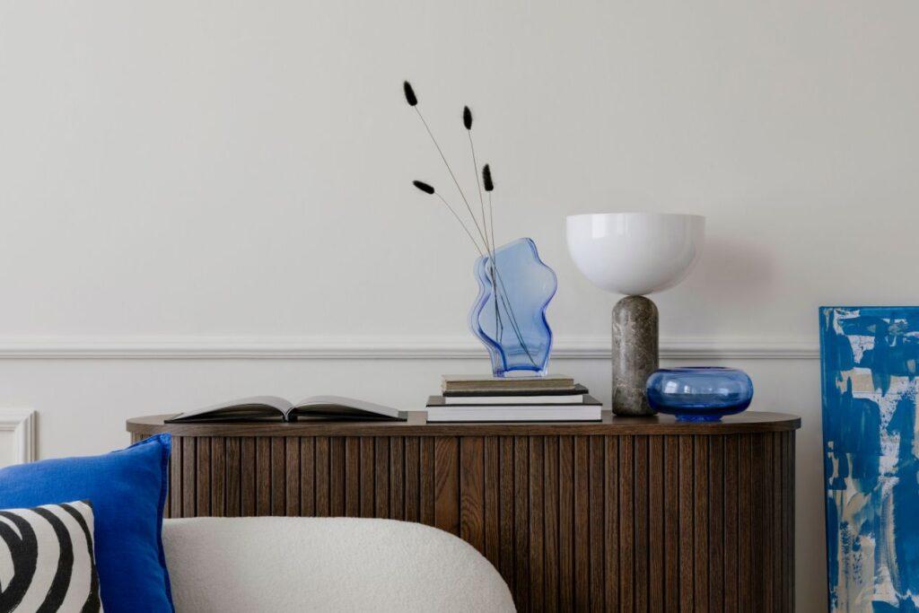 Dunkles Holz-Sideboard mit geriffelter Front, dekoriert mit blauen Glasvasen, Büchern und einer Design-Tischlampe vor einer weißen Wand